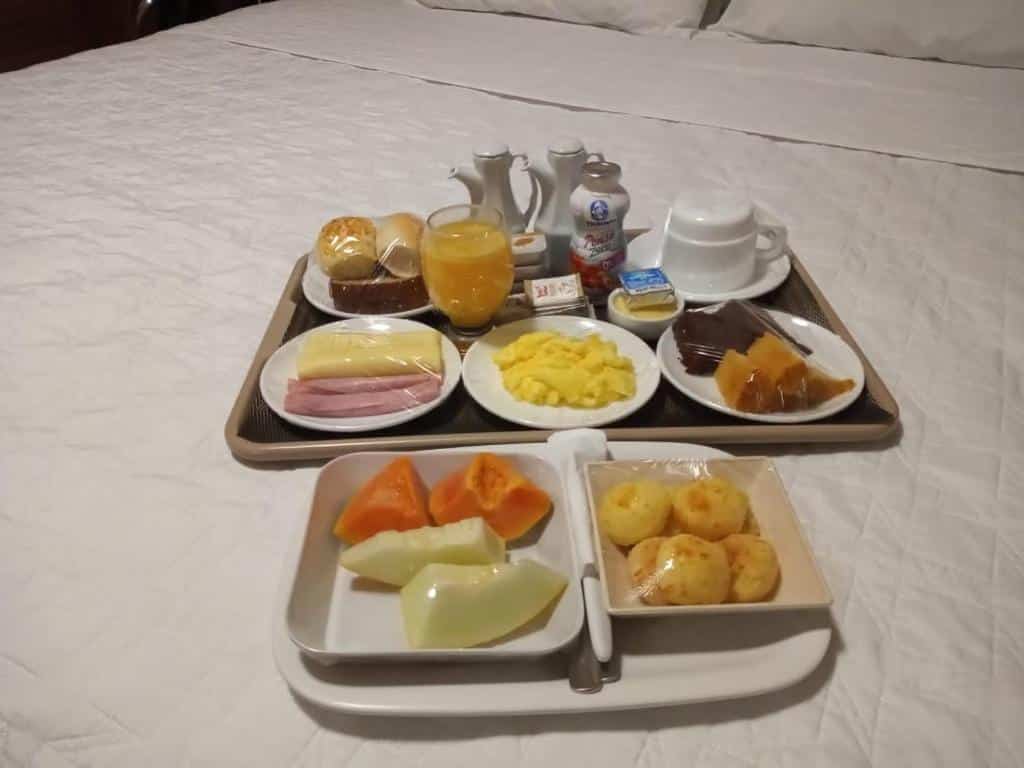 Café na Pousada O Garimpo. Uma bandeja de café da manhã na cama. Frutas, frios, suco, leite, pães, café e frutas.
