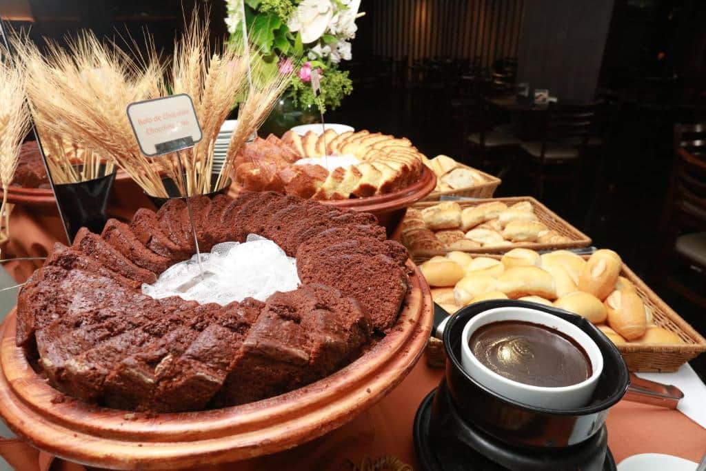 Mesa do café da manhã. Um bolo de chocolate na frente e atrás pães e outros bolos. Foto para ilustrar post sobre Hotéis perto do Hospital Sírio Libanês.