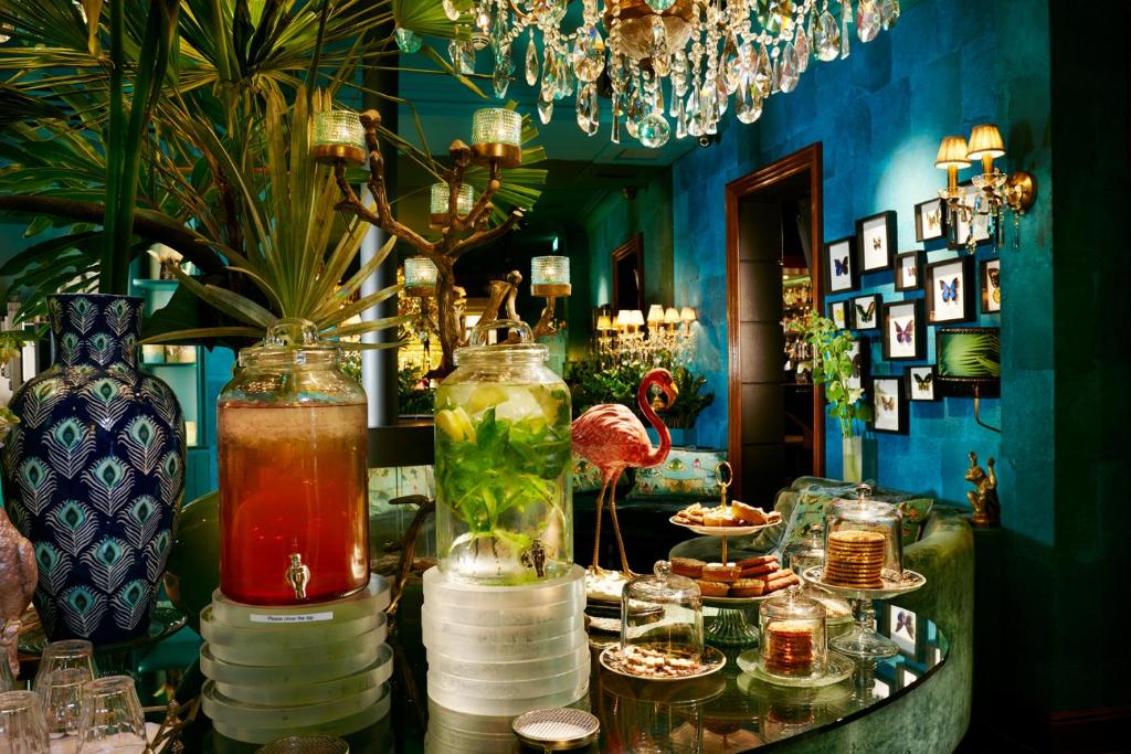 Mesa de vidro do Hotel Estherea, com duas jarras de suco e alguns alimentos de café da tarde. Há plantas decorando o ambiente, que tem parede de cor azul turquesa escuro, lustre, luminárias clássicas e quadros