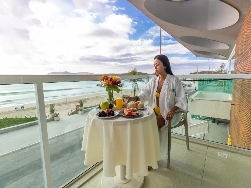 Mulher sentada em uma cadeira em frente a uma mesa com toalha branca, flores e um café da manhã completo. A varanda tem vista para o mar.