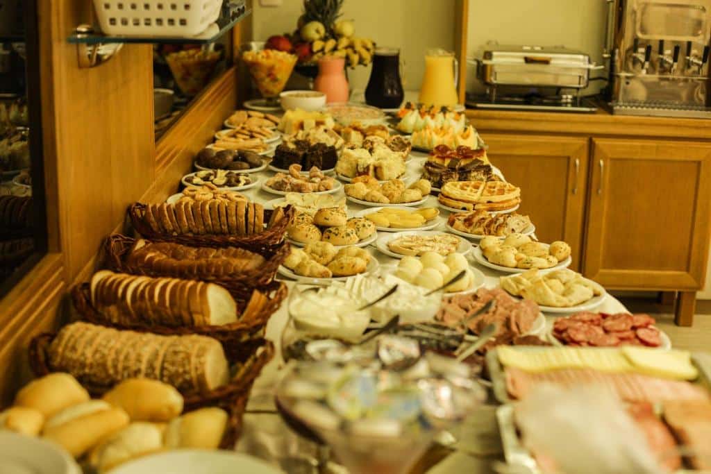 mesa farta de café da manhã com diversos pães, bolos, frios, doces, quitutes e sucos.