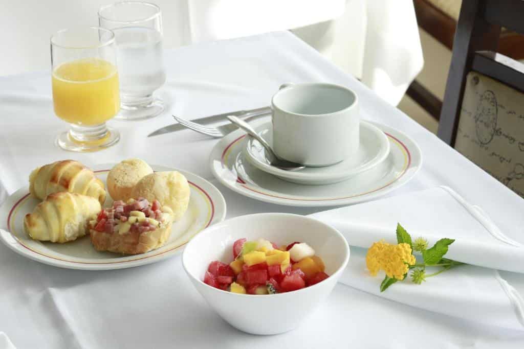 Mesa com café da manhã de hotel com toalha e pratos brancos com salgados e salada de frutas, copos com suco, água e xícara de café.