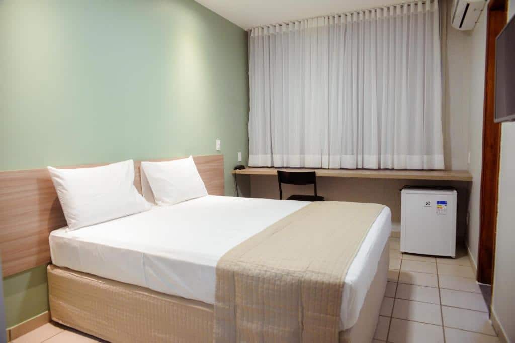 Quarto de hotel com parede verde claro, cama de casal, mesa com cadeira, frigobar e cortinas brancas. Imagem para ilustrar o post hotéis em Teresina.