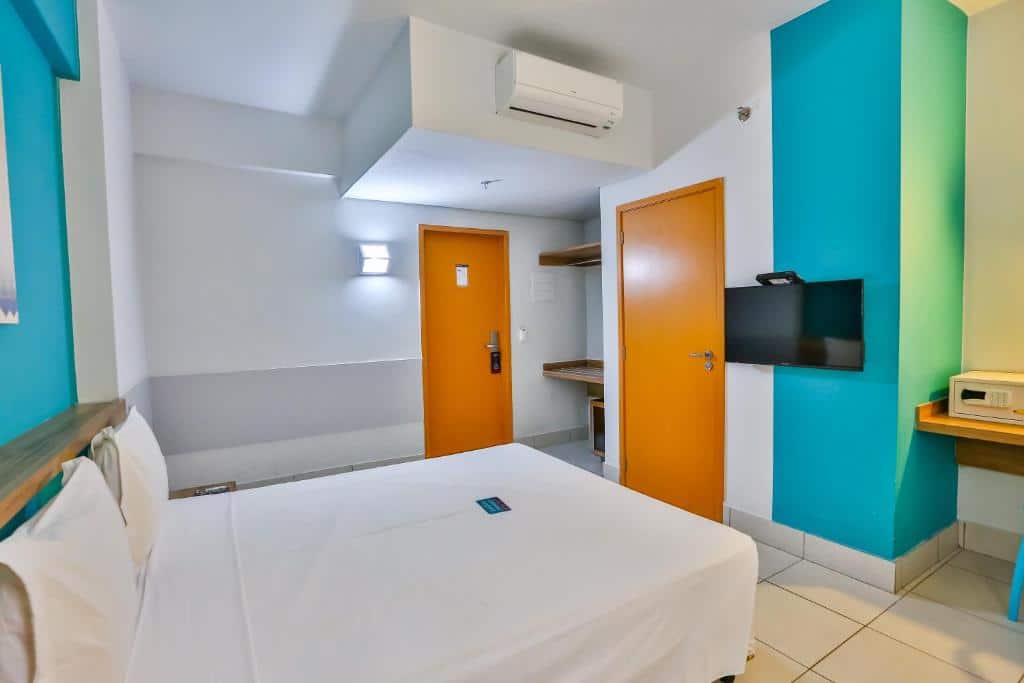 Quarto com paredes branca e detalhes azul, portas de madeira, ar-condicionado, cama de casal com lençóis brancos e TV presa a parede na frente da cama. Imagem para ilustrar o post hotéis em Goiânia.