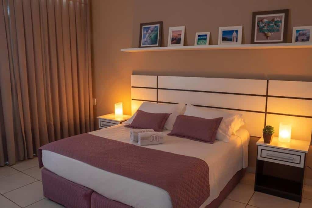 Quarto de hotel com cama de casal, cabeçeira e mesinhas brancas, prateleira com quadros decorativos e cortina. Imagem para ilustrar o post hotéis em Cabo Frio.