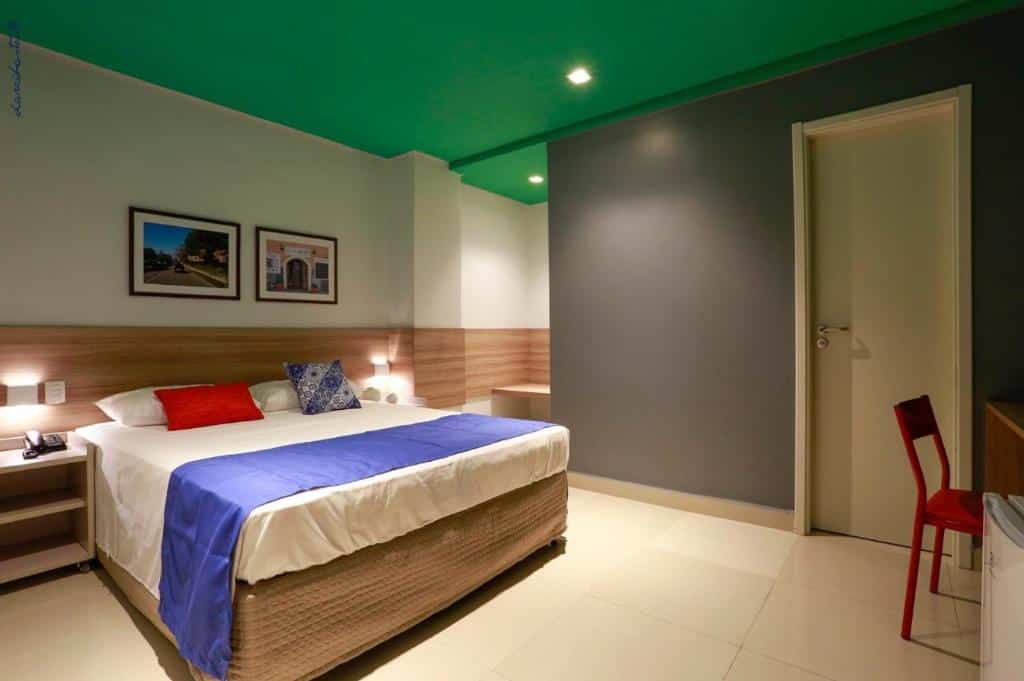 Quarto de hotel com cama de casal, paredes branca, cinza, teto verde, pequenos quadros decorativos, decorações de madeira e luminárias aos lados da cama. Imagem para ilustrar o post hotéis em Teresina.