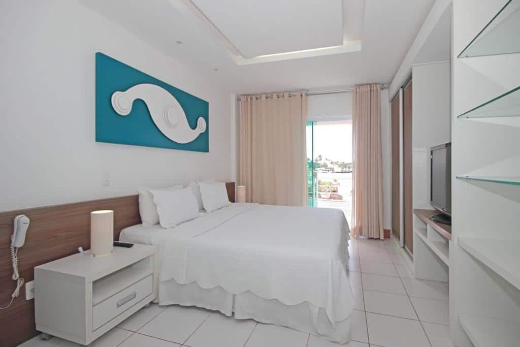 Quarto de hotel com cama de casal, cortinas beje, móveis brancos, televisão, quadro azul e varanda. Imagem para ilustrar o post hotéis em Cabo Frio.