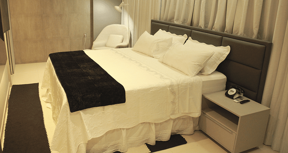 Quarto de hotel luxuoso com grande cama de casal com colcha branca e preta, cabeceira e cortinas cinzas, poltrona branca e tapete cinza escuro. Imagem para ilustrar o post hotéis em Teresina.