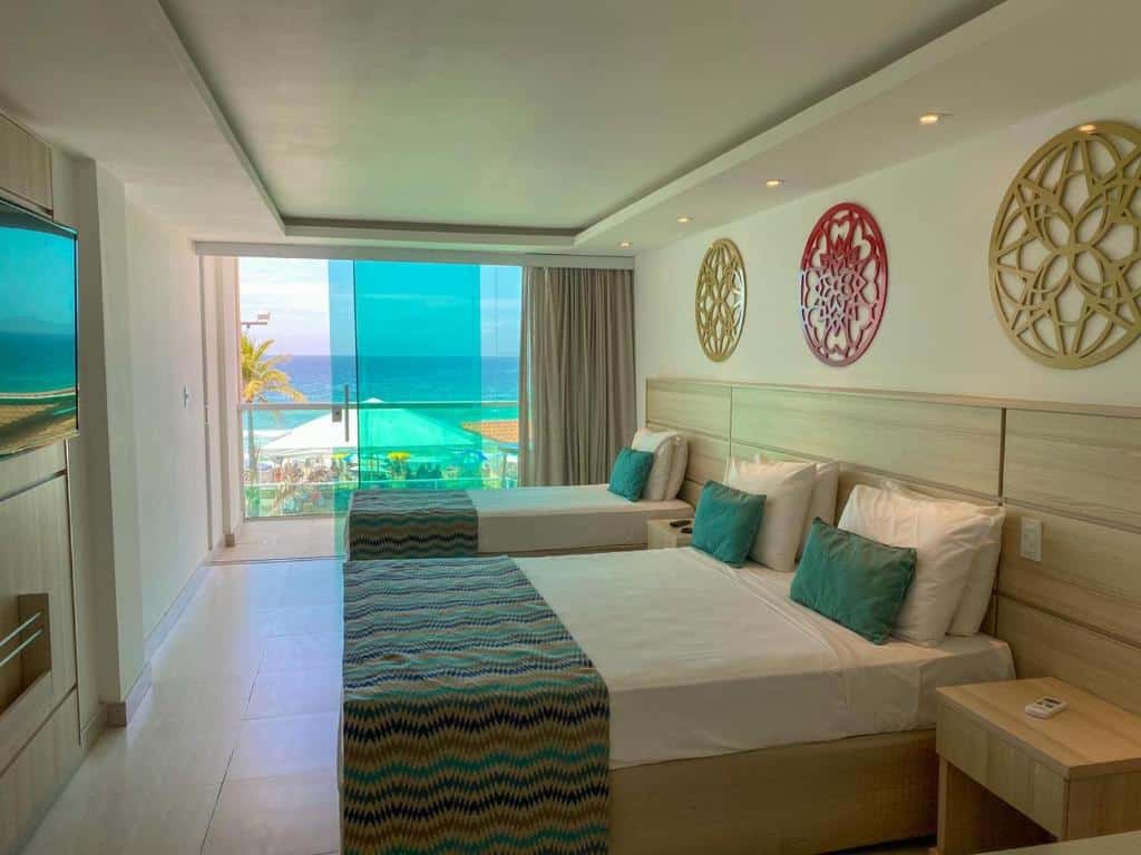Quarto de hotel com uma casa de casal, uma casa de solteiro, cortinas beje, móveis em tons de marrom claro e varanda com vista para o mar. Imagem para ilustrar o post hotéis em Cabo Frio.