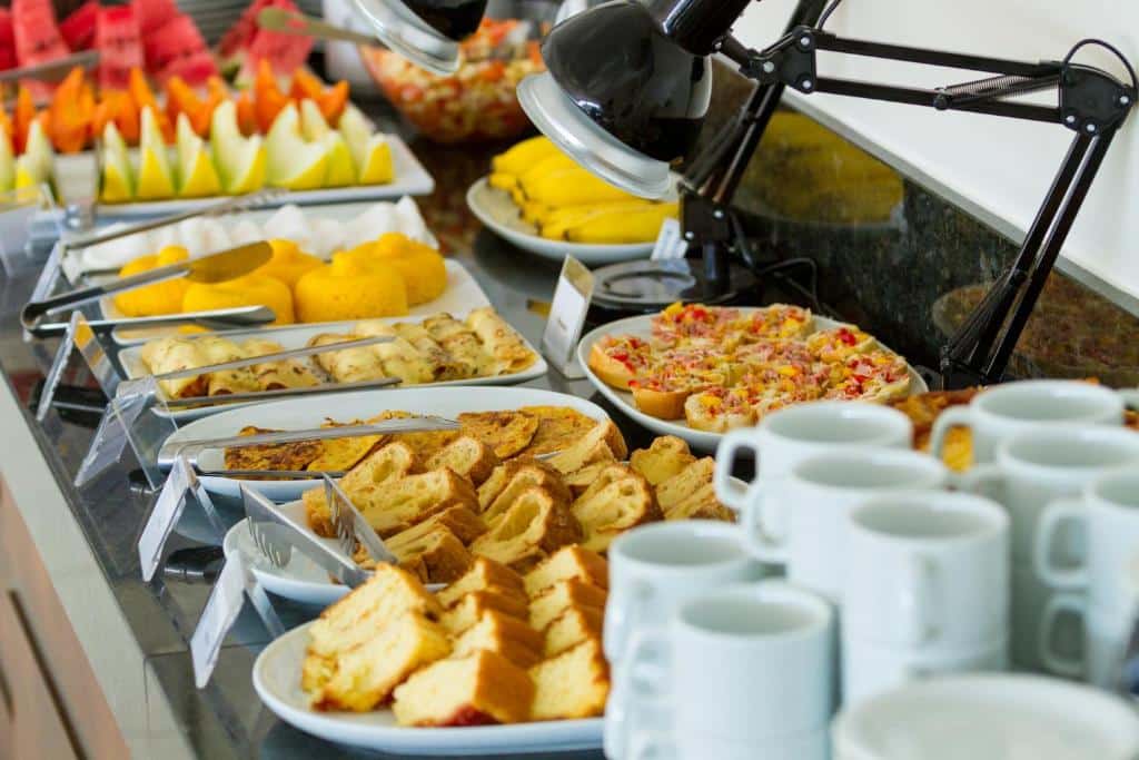 Café da manhã de hotel com canecas a frente, luminárias e pratos brancos com salgados, bolos e frutas.