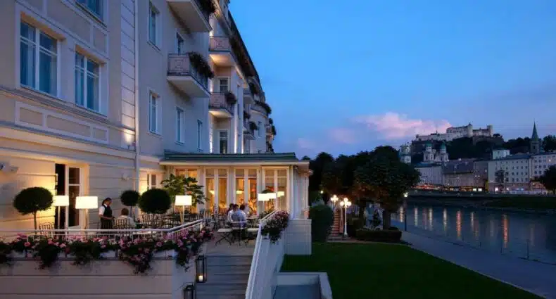 Parte de fora do hotel com algumas pessoas sentadas em cadeiras com vista para o rio e algumas construções antigas da cidade durante o anoitecer, ilustrando post Hotéis em Salzburg.