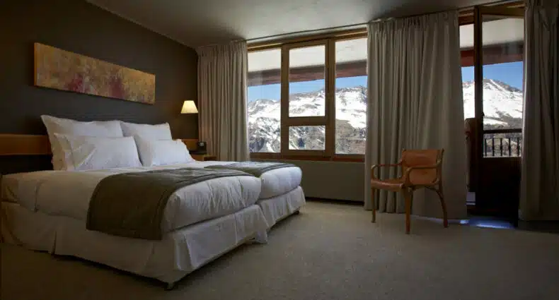Quarto do Valle Nevado com cama do lado esquerdo, uma cadeira de madeira em frente a cama ao lado de uma janela. Representa hotéis no Valle Nevado.