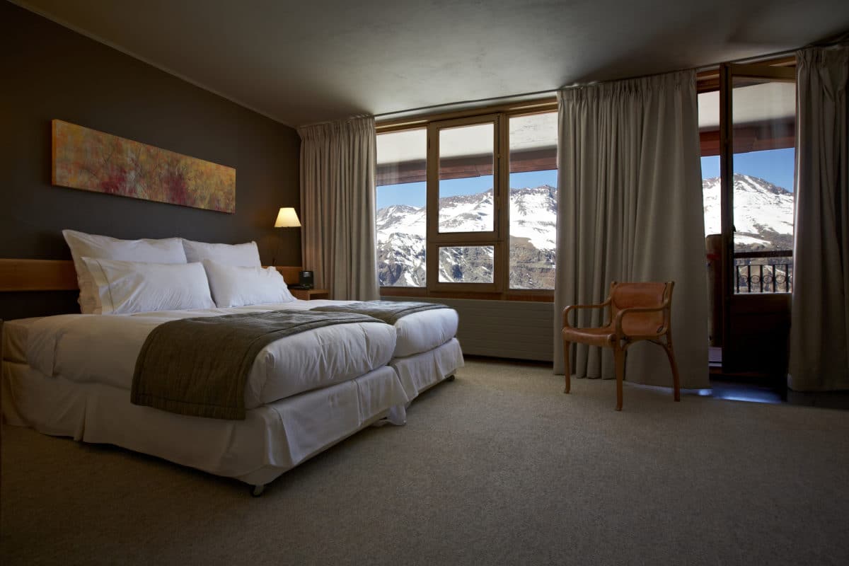 Quarto do Valle Nevado com cama do lado esquerdo, uma cadeira de madeira em frente a cama ao lado de uma janela. Representa hotéis no Valle Nevado.