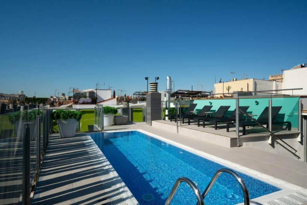 Piscina do Catalonia Born. Esprguiçadeiras pretas encaram a piscina do lado de uma escadaria que leva ao local. Uma grade de vidro protege a área.