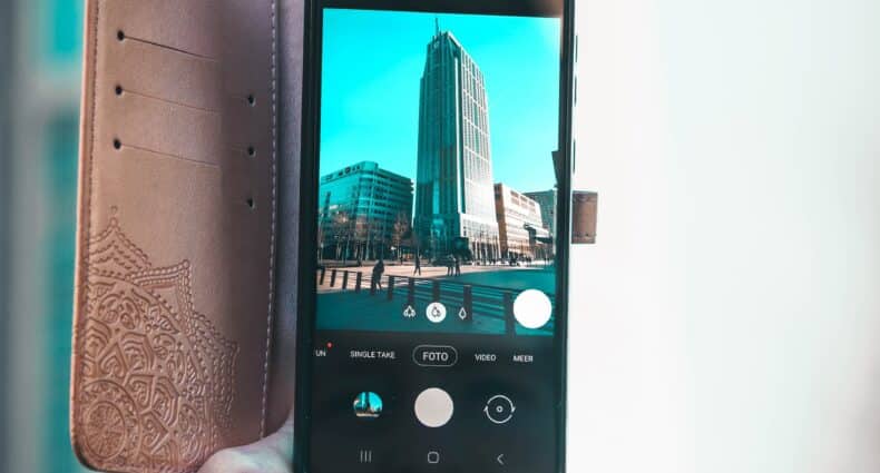 Mão segurando um celular com capinha aberta, câmera posicionada de modo a tirar foto de um prédio, para ilustrar a capa do post sobre chip de viagem