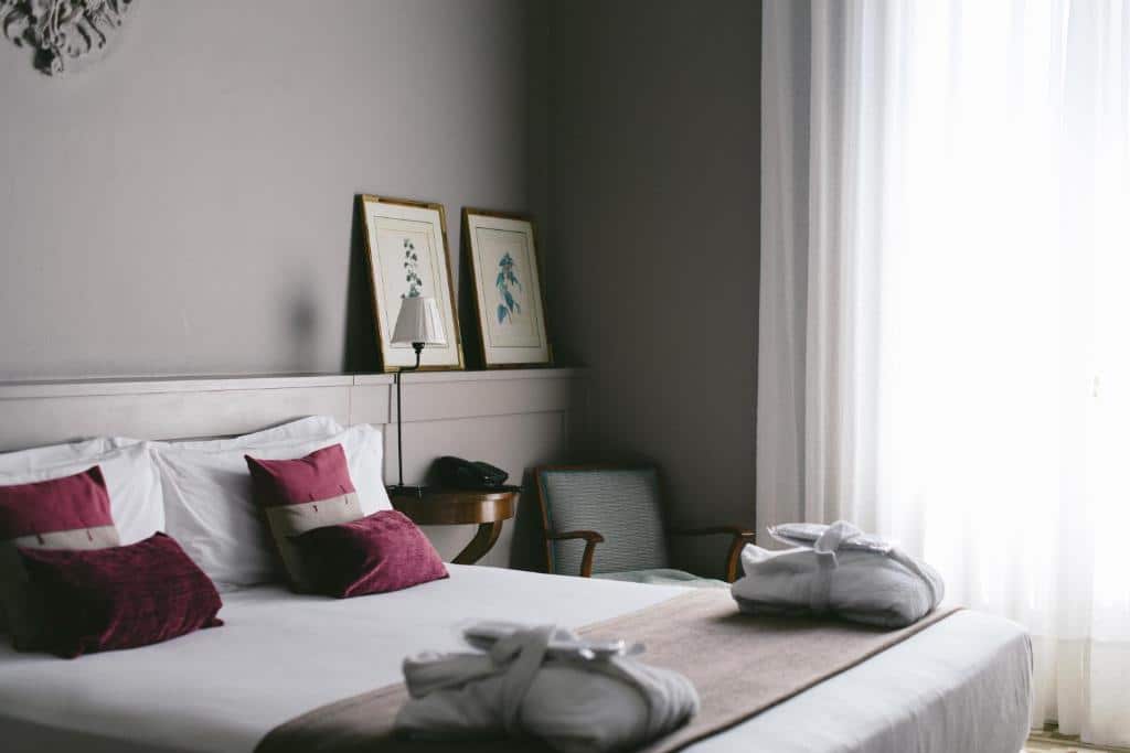 Quarto do Circa 1905, uma das recomendações de hotéis boutique em Barcelona. A cama de casal tem mesinhas de cabeceira com abajur dos dois ado, e uma poltrona está encostada na parede ao lado de uma janela com cortinas brancas.