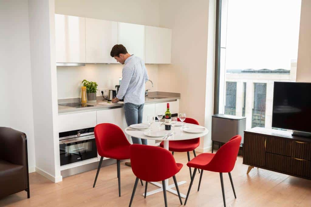 Cozinha de um dos estúdios do Premier Suítes, com uma mesa redonda no meio com 4 cadeiras vermelhas, e um balcão com pia, forno, armário suspenso, e um homem de costas mexendo nos utensílios da cozinha