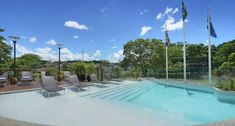Grande piscina em área externa com várias cadeiras ao lado. Ao fundo é possível ver árvores e bandeiras. Foto para ilustrar post sobre hotéis em Campo Grande.