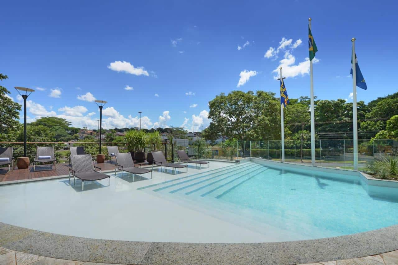 Grande piscina em área externa com várias cadeiras ao lado. Ao fundo é possível ver árvores e bandeiras. Foto para ilustrar post sobre hotéis em Campo Grande.