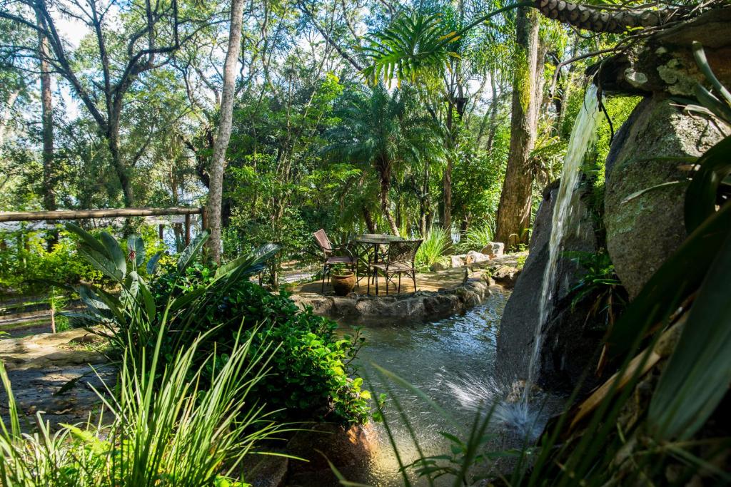 Jardim do Ecoresort Refúgio Cheiro de Mato. Uma bica de água no lado direito no meio das pedras. Atrás do laguinho uma mesa com duas cadeiras. Várias plantas e árvores ao redor.