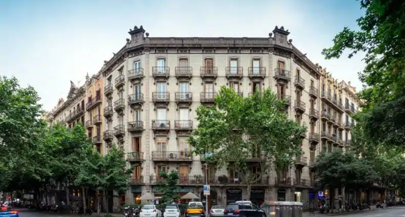 Fachada do Primavera Hostel, uma das recomendações de hostels em Barcelona. O belo prédio bege de cinco andares tem algumas árvores em frente, e carros estão estacionados ao redor do local.