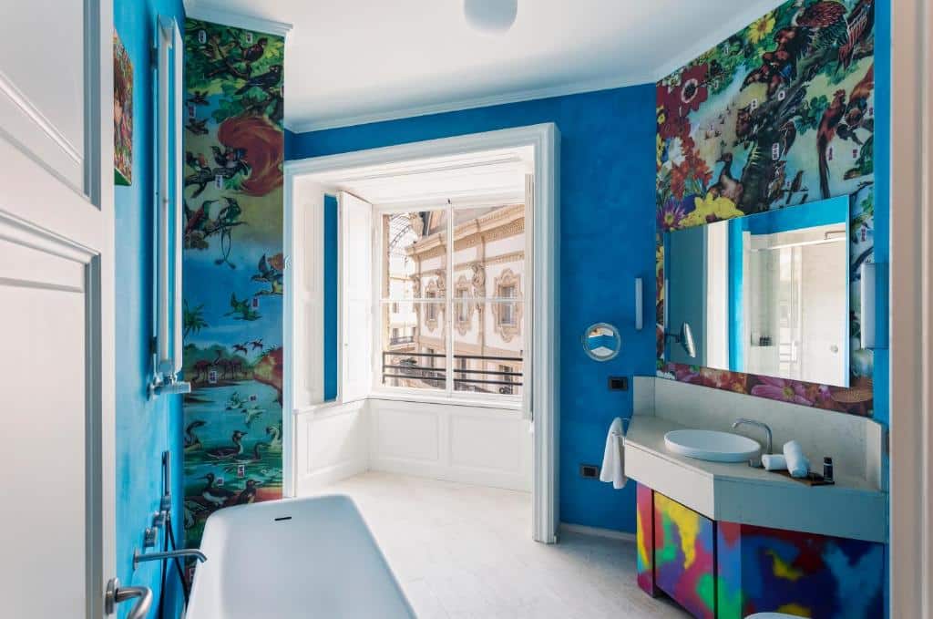 Banheiro decadoro em tons de azul e flores no Galleria Vik Milano com uma janela ampla, um espelho grande, uma pia com toalhas brancas penduradas