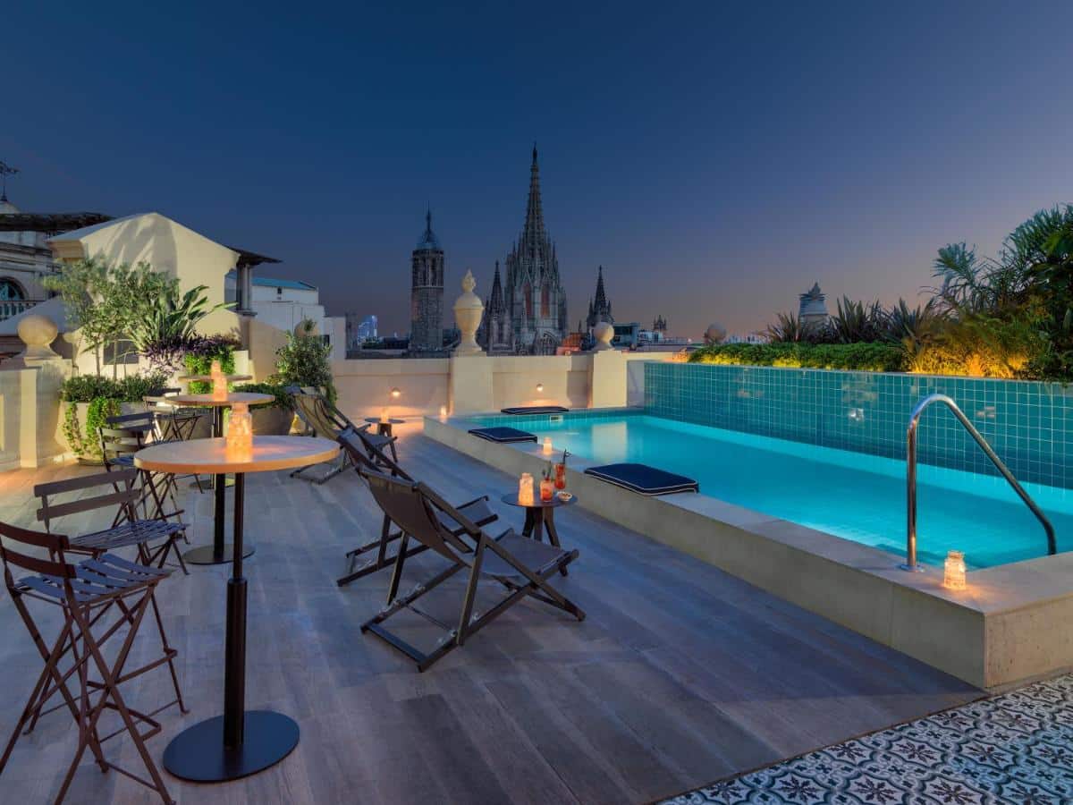 Área da piscina do H10 Madison, uma das recomendações de hotéis no centro de Barcelona. Com vista para a Catedral de Barcelona ao entardecer, a área tem mesas e cadeiras que encaram a piscina. Há escadas e almofadas na beirada da piscina iluminada com luzes aquáticas.