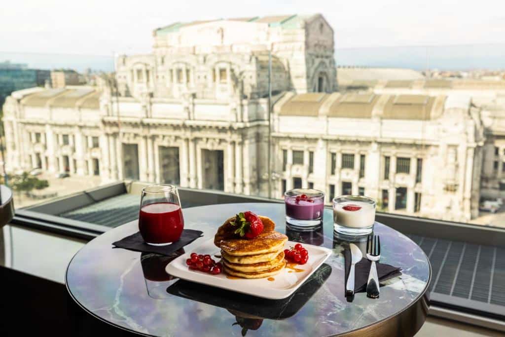 Varanda do HD8 Hotel Milano com uma mesinha redonda com um café da manhã em cima, com panquecas, suco e iogurte, com uma vista para o prédio histórica da Estação Central