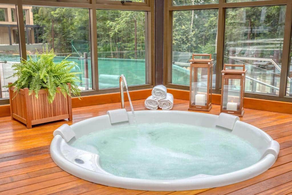 banheira de hidromassagem do Wyndham Gramado Termas Resort & Spa em cima de um deque de madeira com duas lamparinas de vidro ao lado. Atrás é possível ver a piscina e a natureza da região pelos vidros da janela.