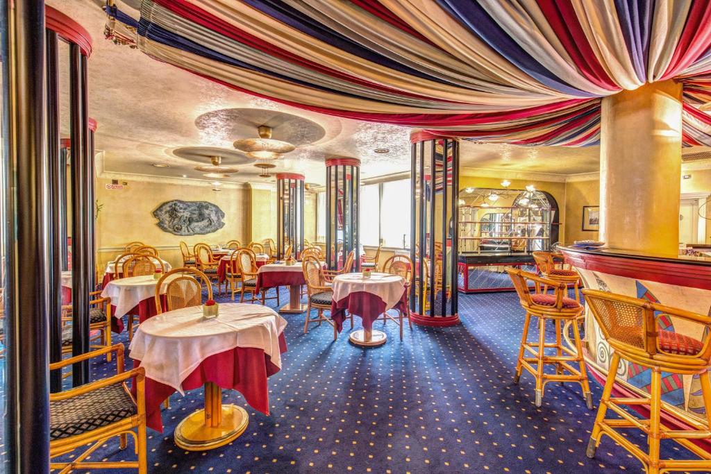 Salão de refeições do Hotel Auriga, o teto é decorado com um pano colorido em branco, azul e vermelho, as mesas com redondas com cadeiras de madeira, o chão tem m carpete azul com pequenos desenhos em dourado, para representar hotéis perto da Estação Central em Milão