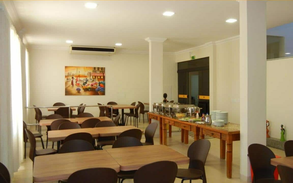 Área de refeição do Hotel Castro com várias mesas e cadeiras, mesa com todas as panelas e pratos e um ar-condicionado no teto.