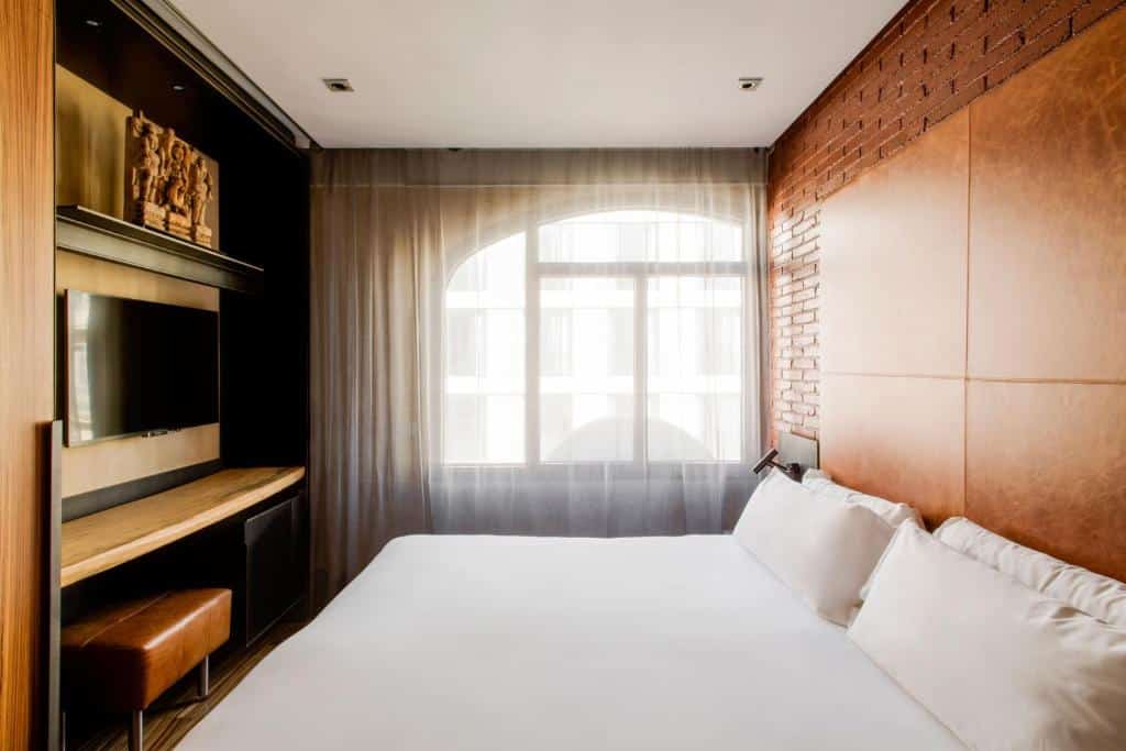 Quarto do Hotel Granados 83, uma das recomendações de hotéis boutique em Barcelona. A cama de casal está encostada na parede do lado direito e encara uma televisão, que tem mesa de trabalho e banco logo abaixo. Ao fundo há uma janela com cortinas bege diáfanas.