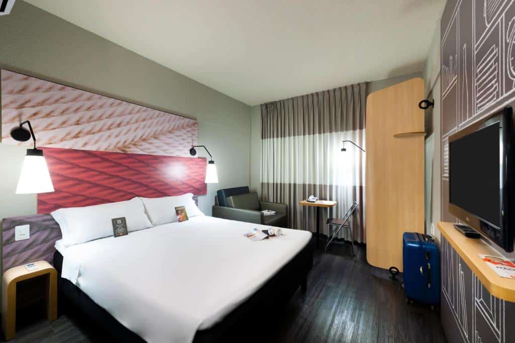 Quarto moderno com cama de casal, luminárias ao lado da cama, poltrona cinza, tv e cortinas. Imagem para ilustrar o post hotéis em Teresina.
