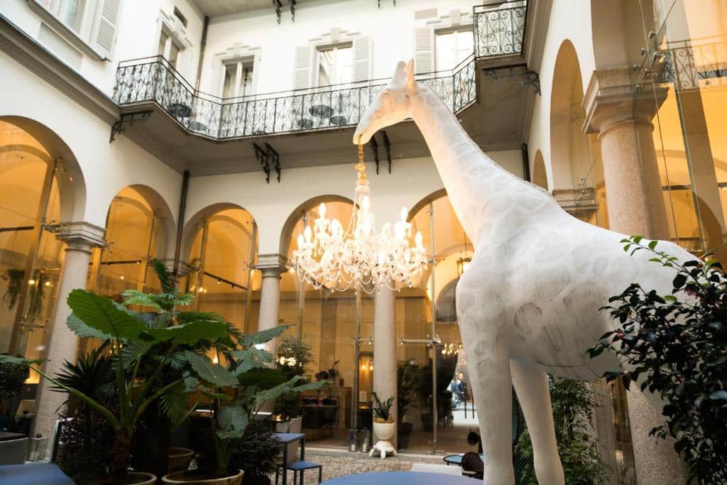 Área aberta do lounge do Hotel Indigo Milan - Corso Monforte, an IHG Hotel com uma escultura de uma girafa branca segurando um lustre na boca, com alguns vasos de plantas, para representar hotéis boutique em Milão