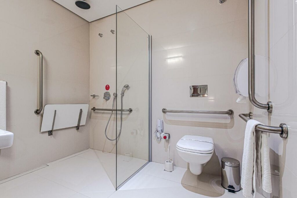 Banheiro com adaptações para PcDs no Hotel Moov Curitiba, com vaso sanitário com barras de apoio, barras no chuveiro e cadeira de assento para o banho, além de pia rebaixada