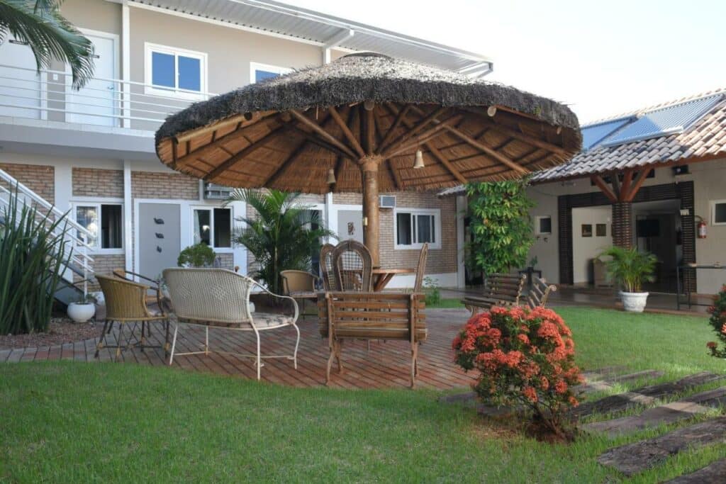 Área externa de uma pousada com grama, flores, mesa, cadeiras e bancos sob um guarda sol. Foto para ilustrar post sobre hotéis em Campo Grande.