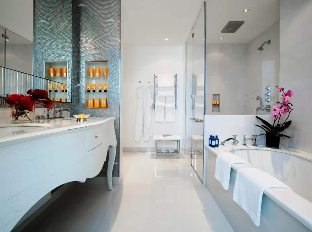 Banheiro do Hotel Principe Di Savoia - Dorchester Collection com uma móvel branco em estilo de época com as cubas da pia, um amplo espelho, um box espaçoso de vidro e uma banheira ao lado, há também roupões e produtos de higiene no local, para representar os melhores hotéis em Milão