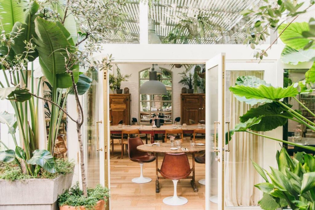 Entrada para o restaurante do Hotel Pulitzer Barcelona. Portas duplas abertas oferecem visão para as mesas e cadeiras do interior do local. Na parte de fora, plantas estão espalhadas entre a fachada branca.