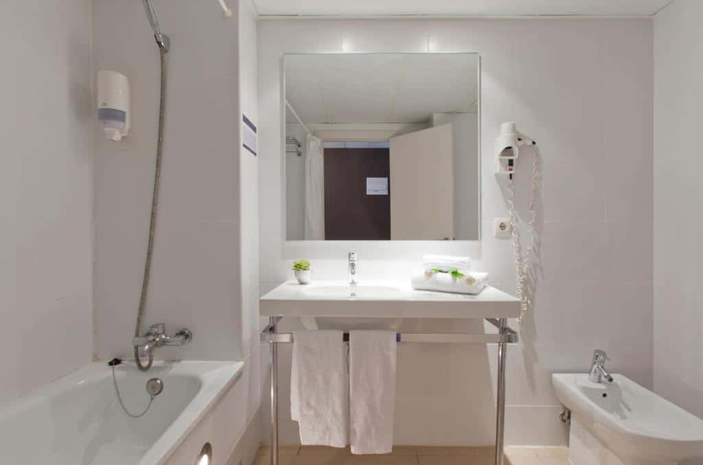 Banheiro do Hotel Sagrada Família. Uma banheira com chuveiro está do lado esquerdo do local. A pia tem espelho, amenidades de banho, toalhas e secador de cabelo e está no meio do banheiro, com uma privada logo ao lado.