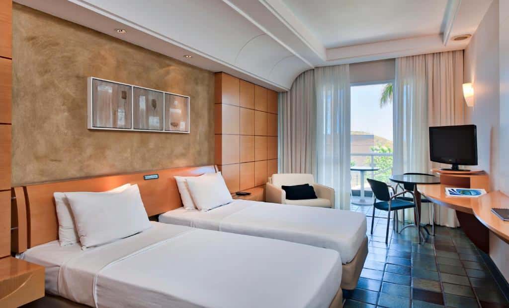 Quarto no Hotel Senac Ilha do Boi com duas camas, mesas e cadeiras, televisão, poltrona e uma janela com cortina branca que leva até a varanda, para representar hotéis em Vitória.