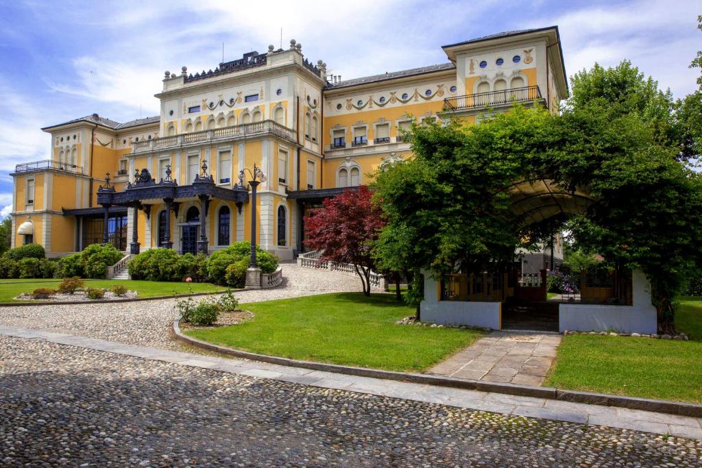Prédio do Hotel Villa Malpensa, a construção é antiga e bem conservada, em tons de branco e amarelo, na frente há muitas árvores e um pequeno gramado, para representar hotéis perto do aeroporto de Milão