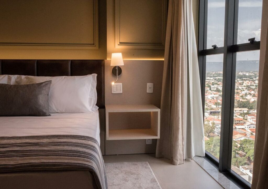 Quarto do hotel Hplus Premium Palmas. Há uma cama de casal, luminária, mesinha e ao lado uma janela grande com cortina e vista para a cidade. Foto para ilustrar post sobre Hotéis em Palmas.