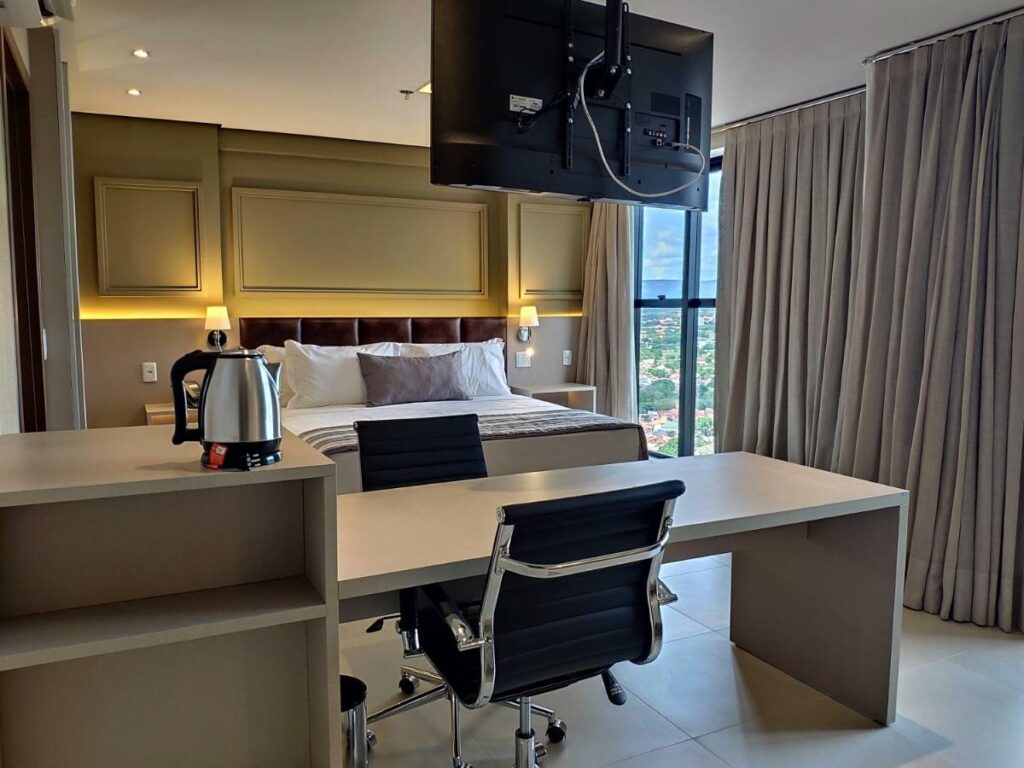 Quarto do hotel Hplus Premium Palmas com uma cama de casal, mesa com cadeira, luminárias, janela grande com cortina e vista da cidade e uma bancada com chaleira. Foto para ilustrar post de hotéis em Palmas.