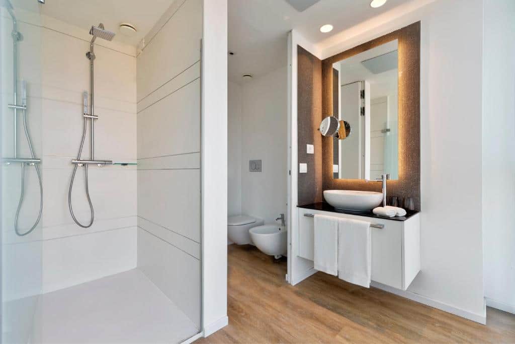 Banheiro do INNSiDE by Meliá Milano Torre GalFa com um box grande, bidê e um vaso sanitário, além de um móvel com toalhas brancas presas, pia e um espelho iluminado