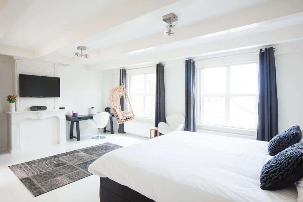 Interior de um quarto do Mokum Suites, com cama de casal, mesa com uma cadeira e, ao lado, uma TV com um móvel abaixo. Há janelas amplas que iluminam bastante o local e um balanço suspenso. Imagem para ilustrar o post hotéis românticos em Amsterdam