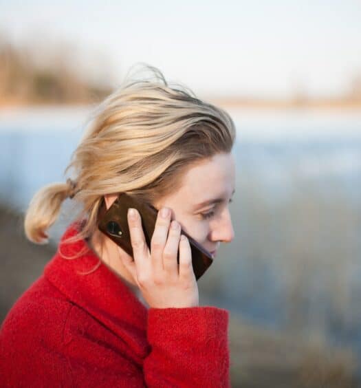 Mulher loira, com casaco vermelho, falando ao celular, com o aparelho posicionado em seu ouvido, tampando parte do rosto. Ao fundo há uma paisagem de lago e vegetação seca