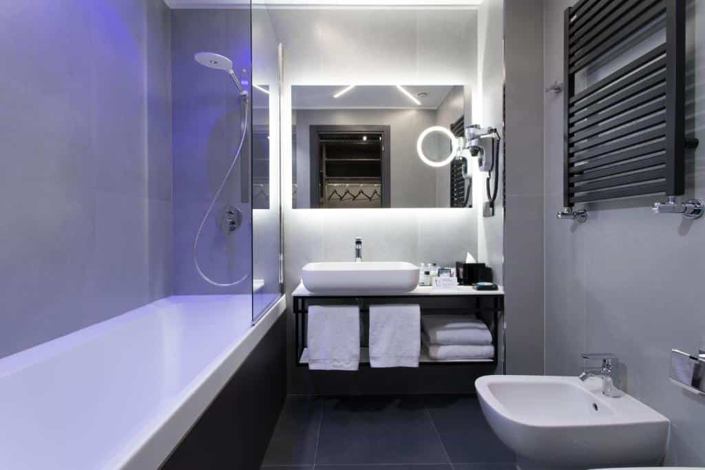 Banheiro do iQ Hotel Milano com uma banheira, um espelho grande, uma cuba com uma pia e toalhas brancas, e um vaso sanitário, para representar hotéis perto da Estação Central em Milão