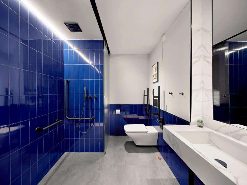 Banheiro adaptado do Kimpton Vividora Hotel. Do lado esquerdo, a parede do chuveiro é de azulejo azul marinho e tem barras de apoio pretas. Do lado direito, uma ampla pia de mármore branco tem um espelho na parede acima. Ao fundo, uma privada branca tem barras de apoio dos dois lados.