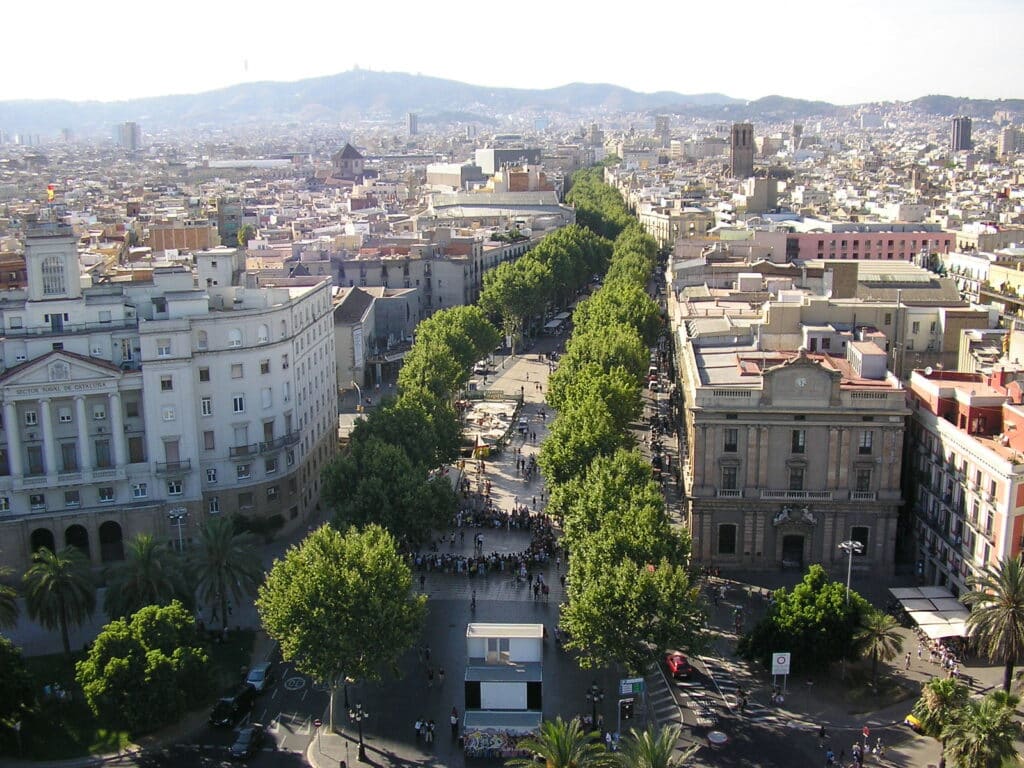 Foto aérea de Las Ramblas. A larga rua tem árvores dos dois lados e muitas pessoas passam por ali. Ao redor é possível ver outras construções de Barcelona. - Foto: Nigel Swales via Flickr