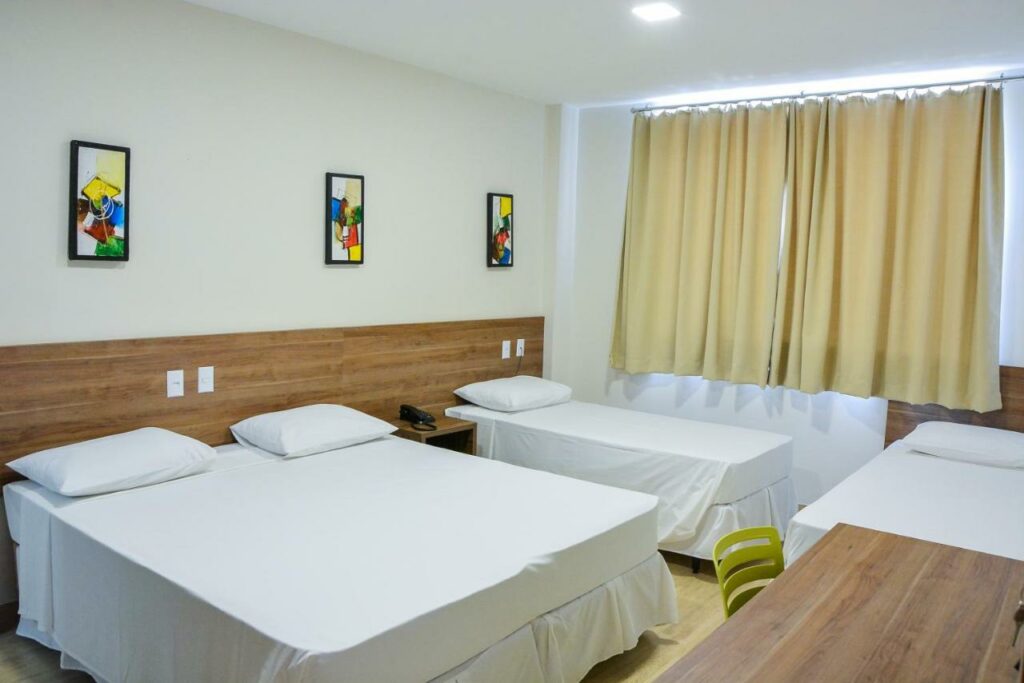 Quarto do Mac Hotel com uma cama de casal e duas camas de solteiro. Há também uma mesa de trabalho com cadeira e uma janela com cortina. Foto para ilustrar post de hotéis em Palmas.